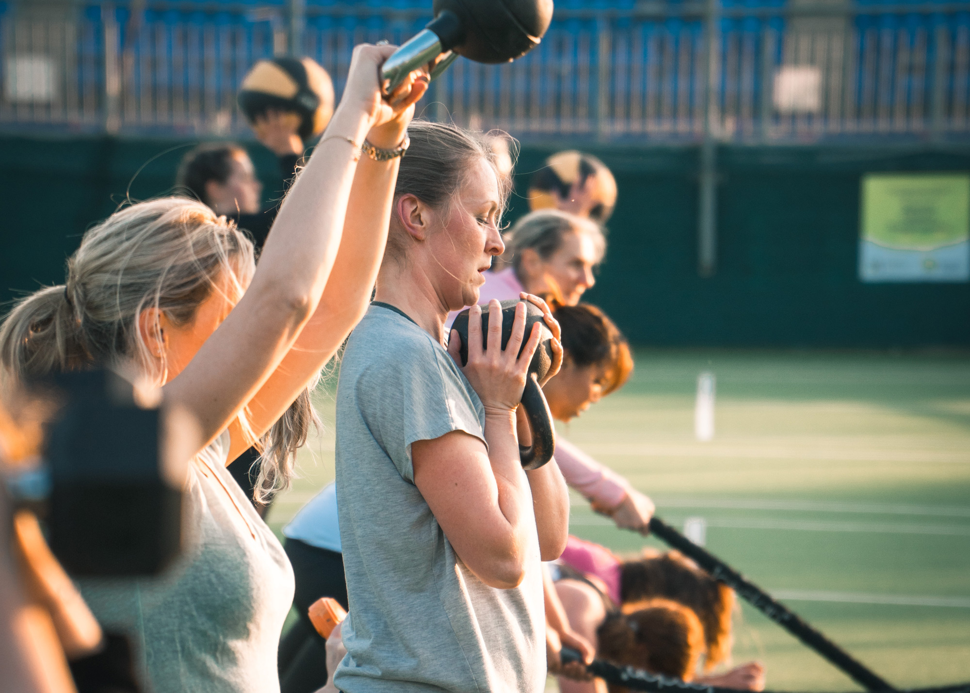 A group session outdoors on the grass, members pressing weights overhead with a coach among them