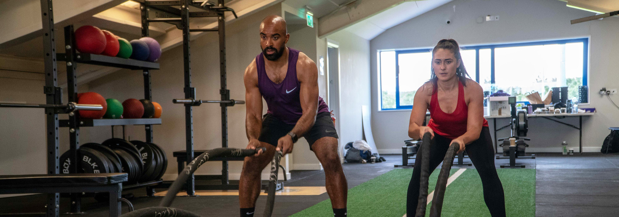 Two members training with weights beside the racks in the studio, daylight through the window behind them